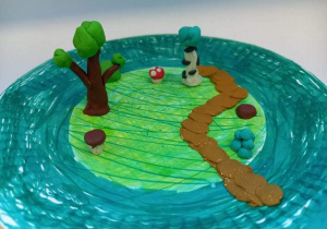 Praca uczestnika zajęć kreatywnych. Papierowy talerzyk pomalowany na zielono. Na talerzyku po lewej stronie stoją dwa drzewka z plasteliny. Między drzewkami stoją dwa grzyby. Po prawej stronie biegnie wyklejona brązowa ścieżka z plasteliny.